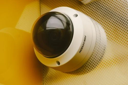 A white Amcrest outdoor security camera mounted on a textured wall, showing the camera lens and housing from a close-up angle. The image illustrates an outdoor security camera setup suitable for monitoring an apartment entrance or exterior area.